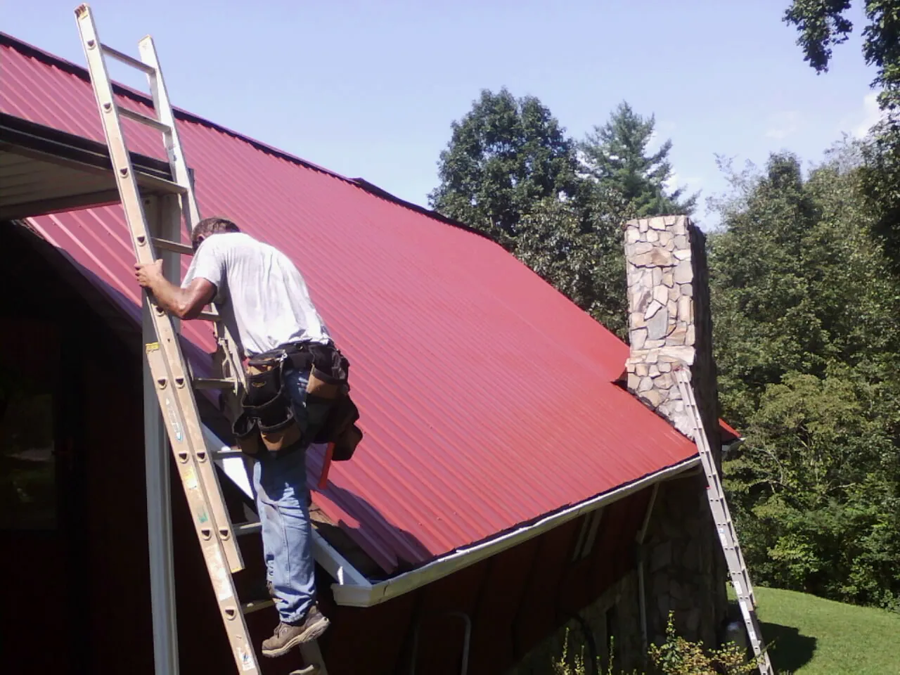 Roofer on a ladder beside a red metal roof and stone chimney — Soffit & Fascia Repair by Roofing Brothers in Hensonville